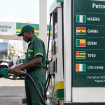 Fuel attendant refuelling a car in Nigeria beside a board comparing August 2026 petrol prices in Nigeria, Ghana, Benin, Togo and Côte d’Ivoire.
